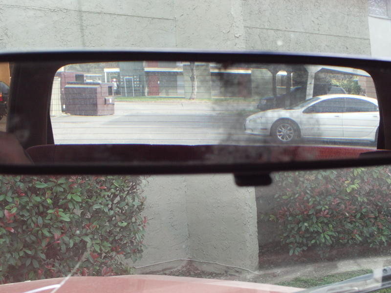 Interior rear view mirror reflection showing a white sedan and a brick structure