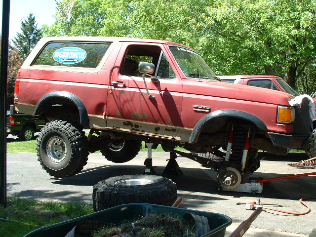 Red Ford Bronco lifted on jack stands with front wheel removed, exposing brake and suspension, in a driveway
