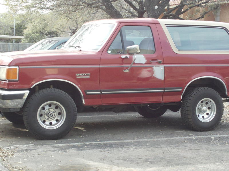 Red Ford Bronco with a tan top, side view, showing body damage on the driver's door