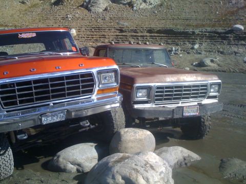 1978 Ford Bronco - Two Ford trucks are parked on a rocky shore with water and large rocks in the foreground.