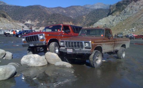 1978 Ford Bronco - Two classic Ford trucks are shown off-road in a rocky, watery area with mountains in the background.