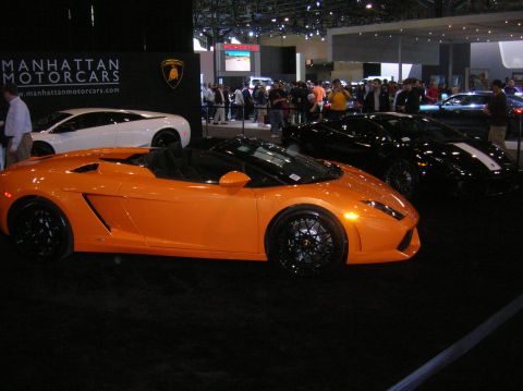 1996 Ford Bronco - An orange convertible sports car is displayed at an auto show next to other vehicles.