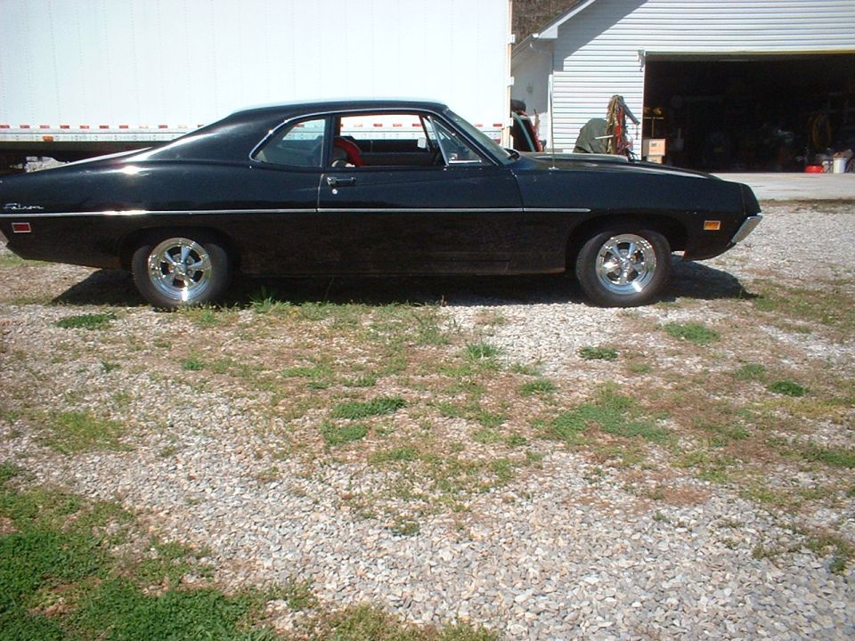 Black two-door coupe in side profile parked on gravel near a trailer and shed.