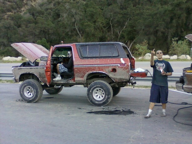 Red 1978 Ford Bronco with hood open, driver seated, and a man standing beside it