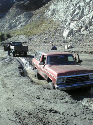 1978 Ford Bronco - A red 1978 Ford Bronco is stuck in a muddy rut, with a pickup truck visible behind it on a dirt road.