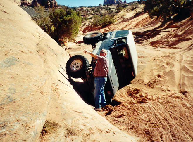 Dark pickup tipped at steep off-camber angle in slickrock gully with man pointing out the line