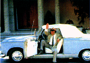 1991 Ford Crown Victoria - A man in a suit waves from the driver's seat of a light blue convertible.