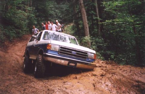 2002 Ford F-350 - A two-tone Ford Bronco tackles a steep, muddy incline in a wooded area.