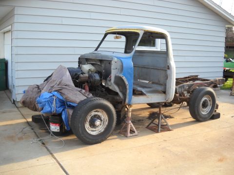 1954 Ford F-100 - A partially disassembled truck sits on jack stands in front of a garage, awaiting restoration.