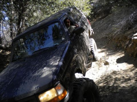 1995 Ford Bronco - A mud-covered SUV navigates a steep, narrow dirt path through a wooded area.