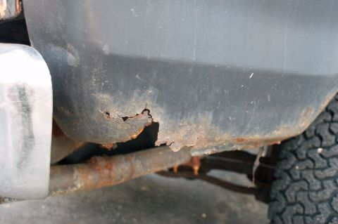 1979 Ford Bronco - Detail of significant rust damage on the lower body panel and exhaust pipe of a vehicle.