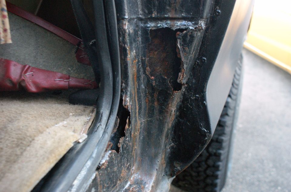Black vehicle door frame with extensive rust damage, showing exposed metal and holes. Interior carpet and red seat upholstery visible