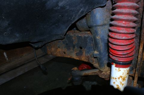 1979 Ford Bronco - Close-up view of heavily rusted vehicle frame and suspension components, including a shock absorber with a red protective boot.