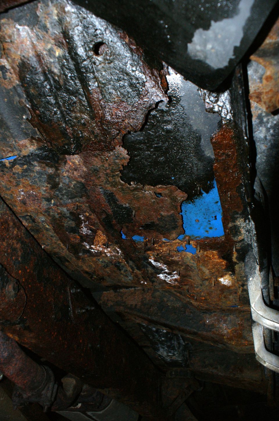 view of heavily rusted frame and undercarriage components, with some blue paint visible through rust holes