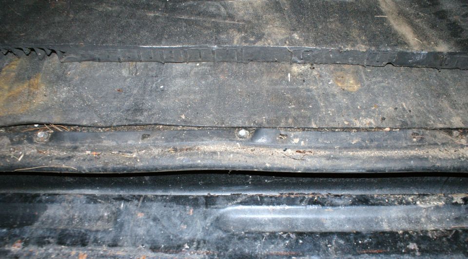 Close-up of the dirty, rusty undercarriage of a vehicle, showing metal panels and some debris