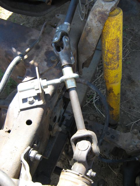 1972 Ford F-250 - Close-up of a steering shaft with two universal joints connecting a black upper shaft through a body-mount support plate down to a rusty lo…
