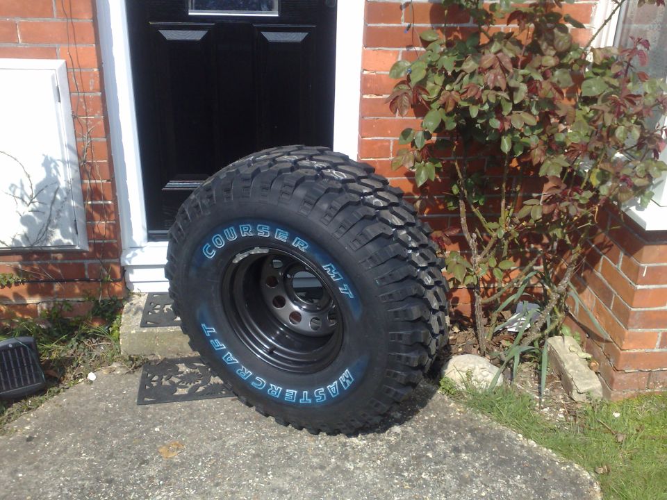 Mud-terrain tire on a black steel wheel by a brick doorway