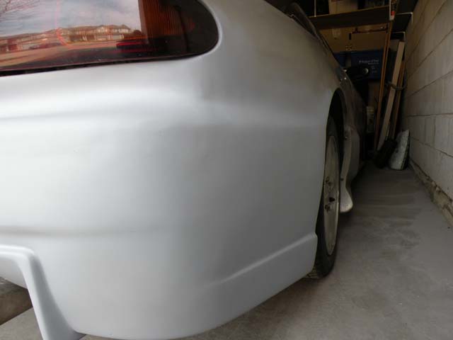 Car body, possibly a coupe, in primer, parked in a garage. Rear quarter panel and wheel visible