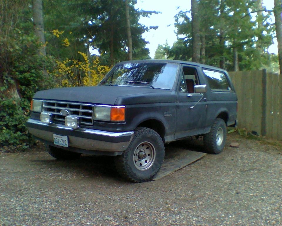 1987 Ford Bronco parked on gravel with trees and fence in background. License plate reads BGT-ZVC