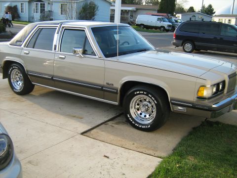 1988 Mercury Grand Marquis - Front three-quarter view of a tan/beige Mercury four-door sedan with a padded vinyl half-roof, chrome window trim and a 5.0 front-fender ba…