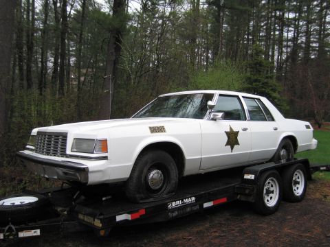 1991 Ford LTD Crown Victoria - A white 1979 Ford LTD Crown Victoria sheriff's car is loaded onto a trailer, showing its front 3/4 view with a flat tire.
