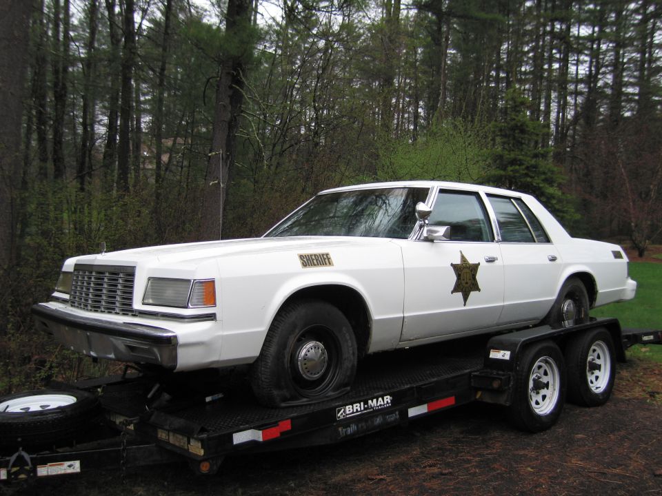 White 1979 Ford LTD Crown Victoria sheriff's car on a trailer, front 3/4 view, flat tire on front passenger side