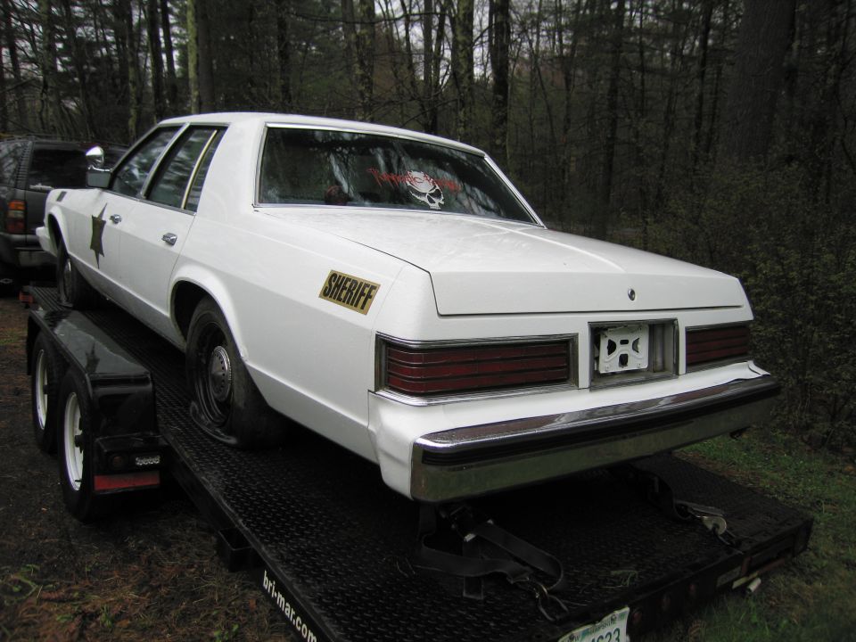 White sedan with a flat rear tire on a trailer, "SHERIFF" decal on the door