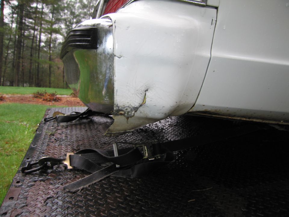 Damaged white rear bumper and quarter panel with chrome trim, secured with straps on a trailer
