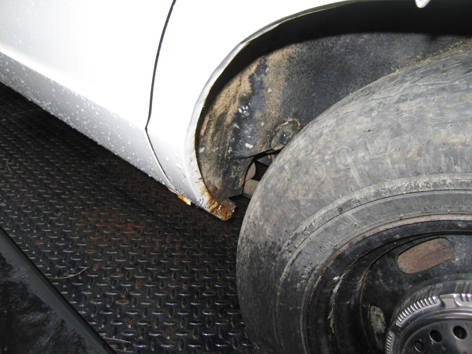 Close-up of a white car's rear wheel well showing rust and a worn tire on a black metal surface