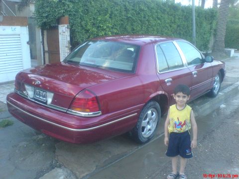 1998 Ford Crown Victoria - A young boy stands beside a maroon sedan parked on a wet street with a hedge in the background.