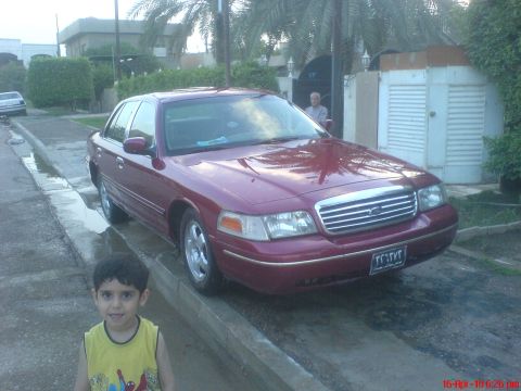 1998 Ford Crown Victoria - A maroon 1998 Ford Crown Victoria is parked on a wet street, with a child in the foreground and a man visible in the background.
