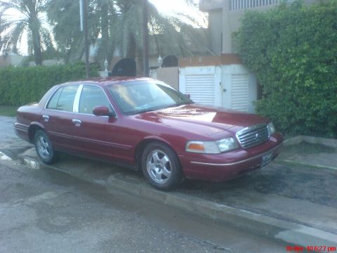 1998 Ford Crown Victoria - A maroon 1998 Ford Crown Victoria is parked on the side of a street with palm trees and a building in the background.