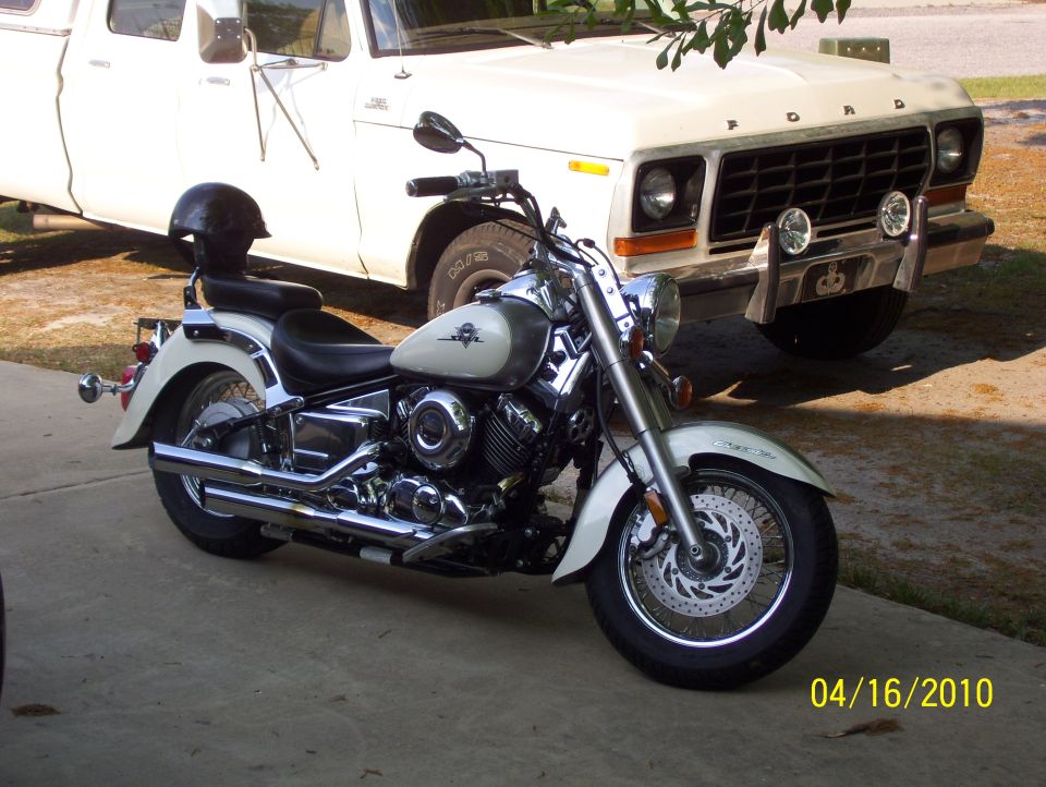 White Ford truck with a chrome grille and auxiliary lights parked behind a cream and silver motorcycle with a black helmet