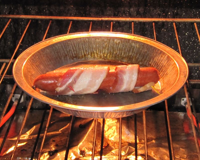 Hot dog wrapped in bacon cooking in a foil pan inside an oven