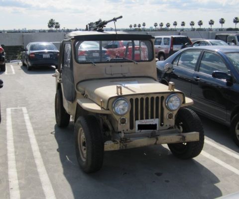 1990 Ford Bronco - A tan jeep, possibly a military surplus vehicle, is parked in a lot with other cars under a partly cloudy sky.