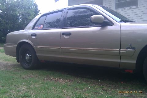2000 Mercury Grand Marquis - Side view of a tan sedan with black wheels and an American flag sticker on the rear window.