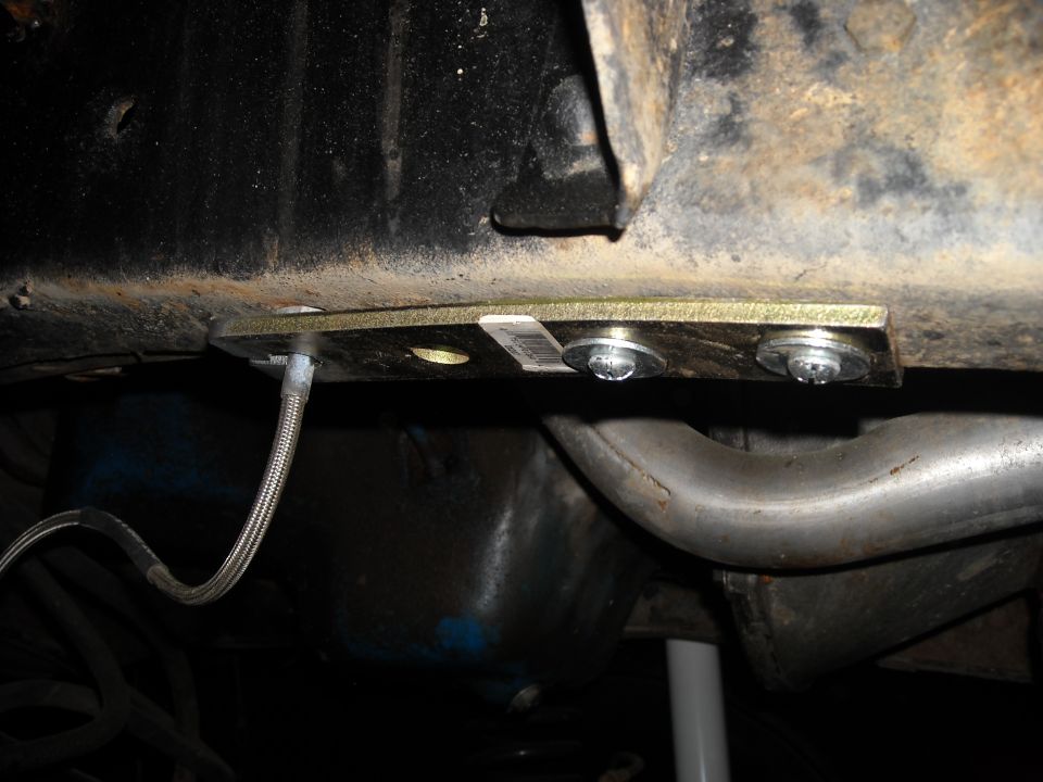 Close-up of a metal plate bolted to a rusty frame rail, with a braided brake line attached. Exhaust pipe visible below