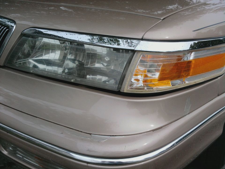 Tan sedan's front corner with chrome trim, clear headlight, and amber turn signal. Scratches visible on bumper