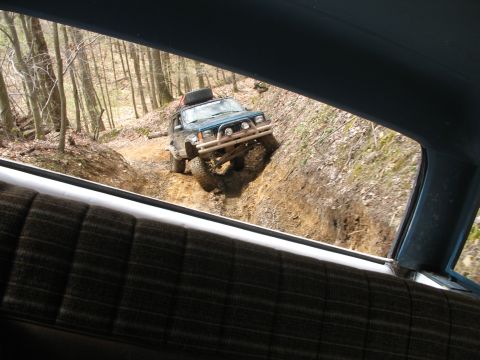 1985 Ford Bronco - A Jeep Cherokee XJ navigates a challenging off-road trail, with the interior of another vehicle visible in the foreground.