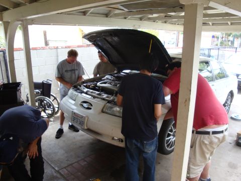 1995 Ford Taurus - A group of people are gathered around the open hood of a white sedan, appearing to work on the engine.