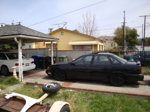 1995 Ford Taurus - A black 1995 Ford Taurus sedan is parked in a driveway next to a white car under a carport.