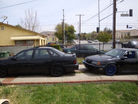 1995 Ford Taurus - Two black sedans are parked next to each other on a sidewalk in front of a house.