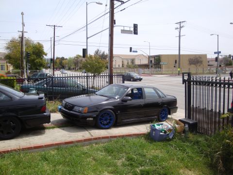 1995 Ford Taurus - A black sedan with custom blue rims is parked on a sidewalk, with another vehicle partially visible.