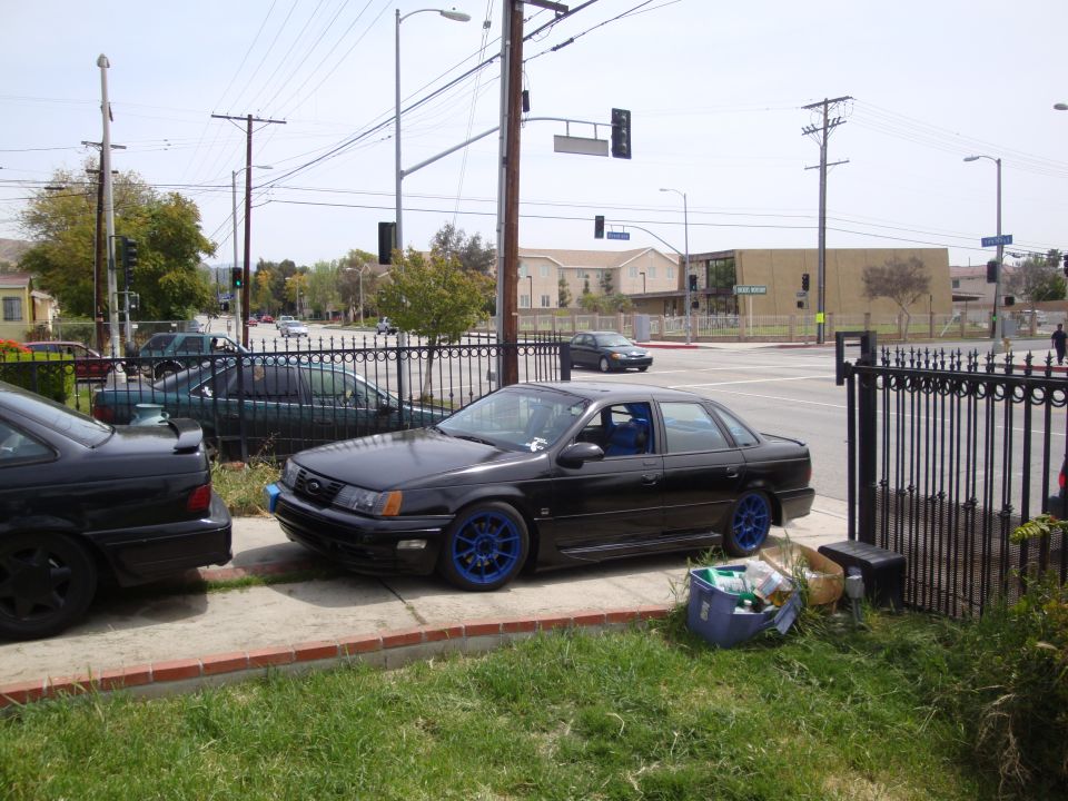 Black sedan with blue rims parked on sidewalk next to another car. Trash bin nearby