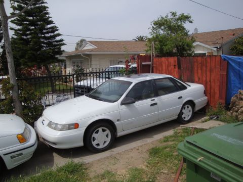 1995 Ford Taurus - A white sedan is parked in a driveway next to another vehicle, with a house and fence in the background.