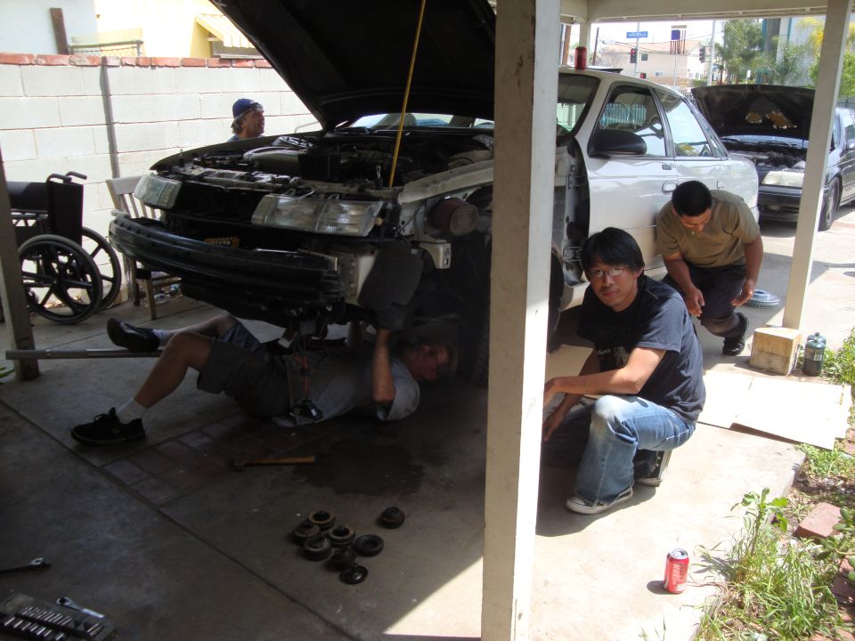 Three people working on the front end of a car, with another car visible behind it