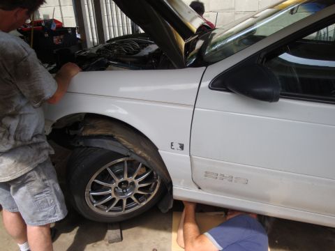1995 Ford Taurus - Mechanics are working on the front wheel and engine bay of a white sedan.