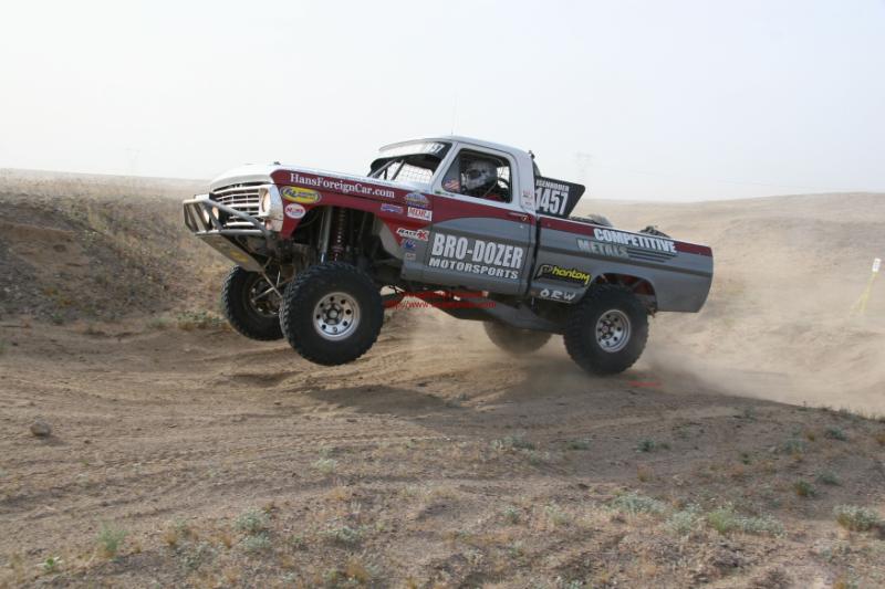 Custom off-road truck mid-air jump, red and gray paint, 'BRO-DOZER MOTORSPORTS' and 'COMPETITIVE METALS' decals visible