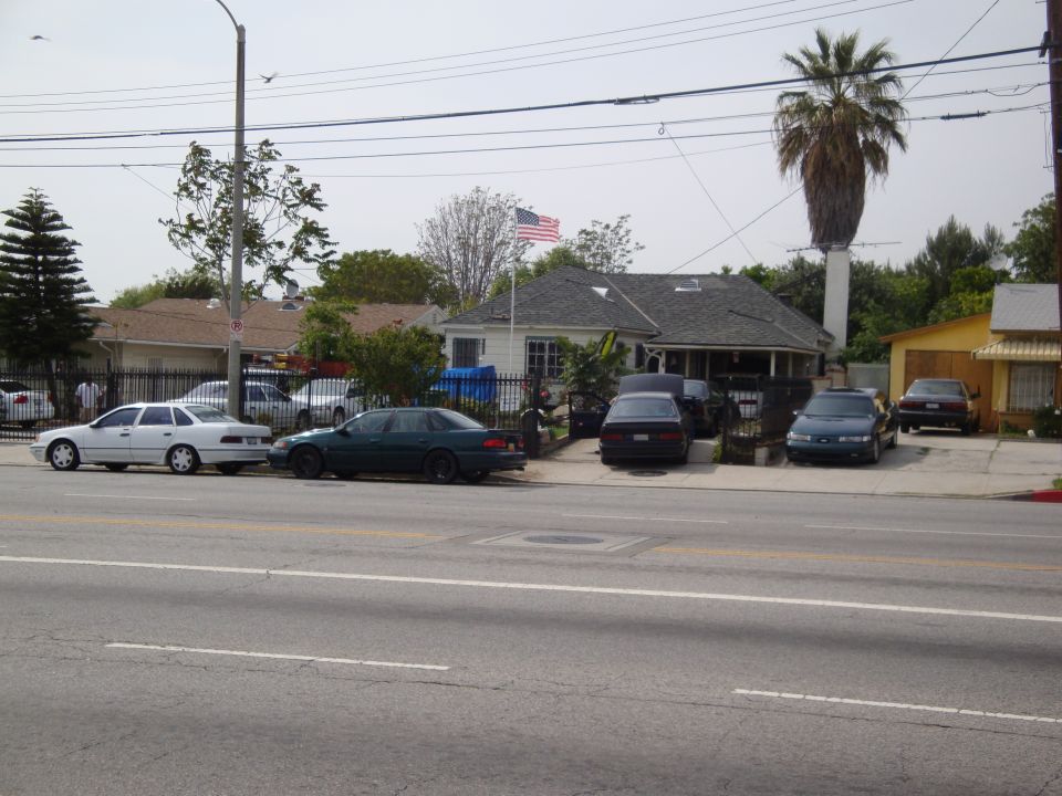 Several cars parked on a street in front of houses. One white sedan is on the left, a dark green sedan is next to it. More cars are parked in driveways