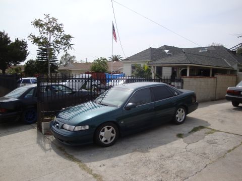 1995 Ford Taurus - A dark green 1995 Ford Taurus is parked in a driveway next to another vehicle.